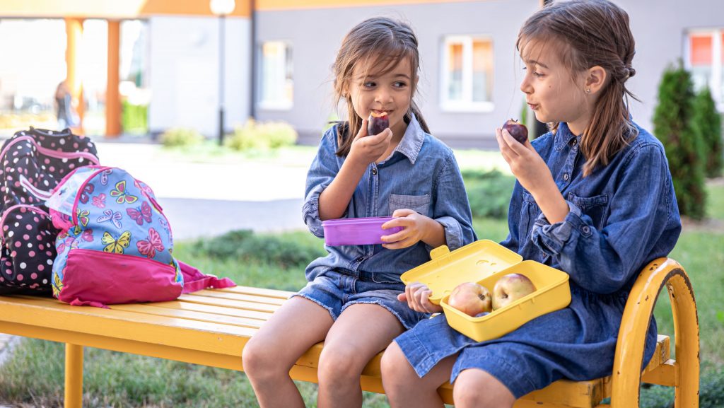 Little school girls sitting on bench in school yard and eating fruits outdoor.