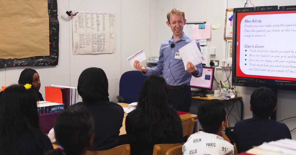Specialist Music PPA Cover Teacher laughing, holding resources ready to deliver a fun music lesson to a classroom of primary school pupils.